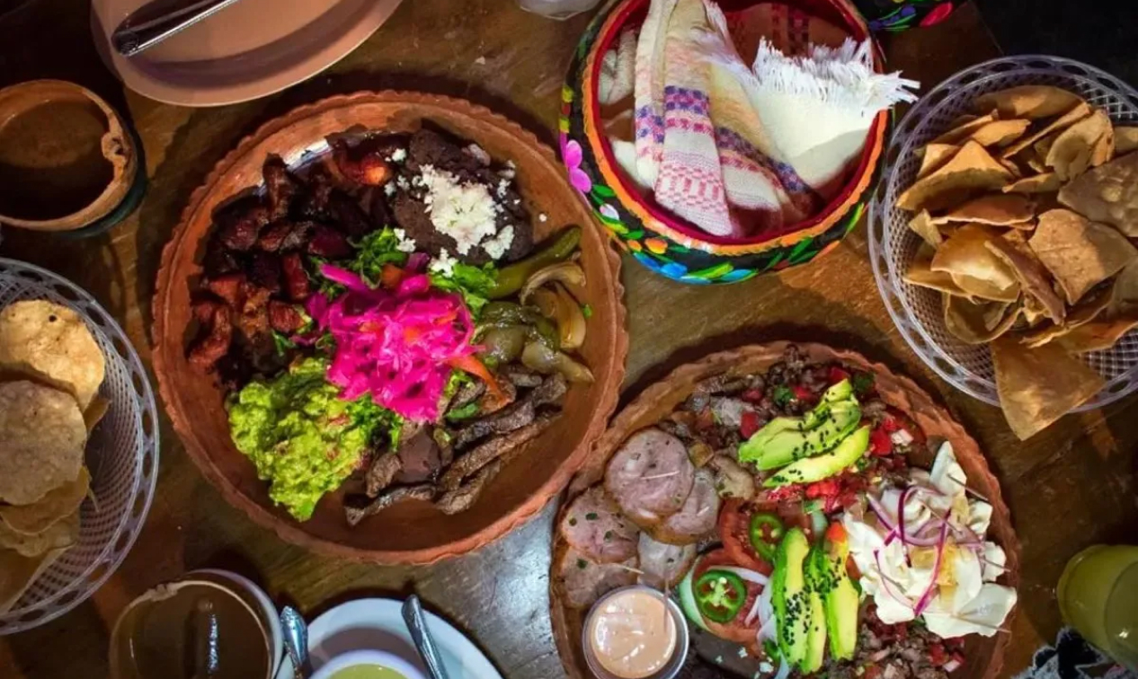 Grilled meats, black beans, guacamole, pickled onions and warm tortillas arranged on terracotta platters across a wooden table